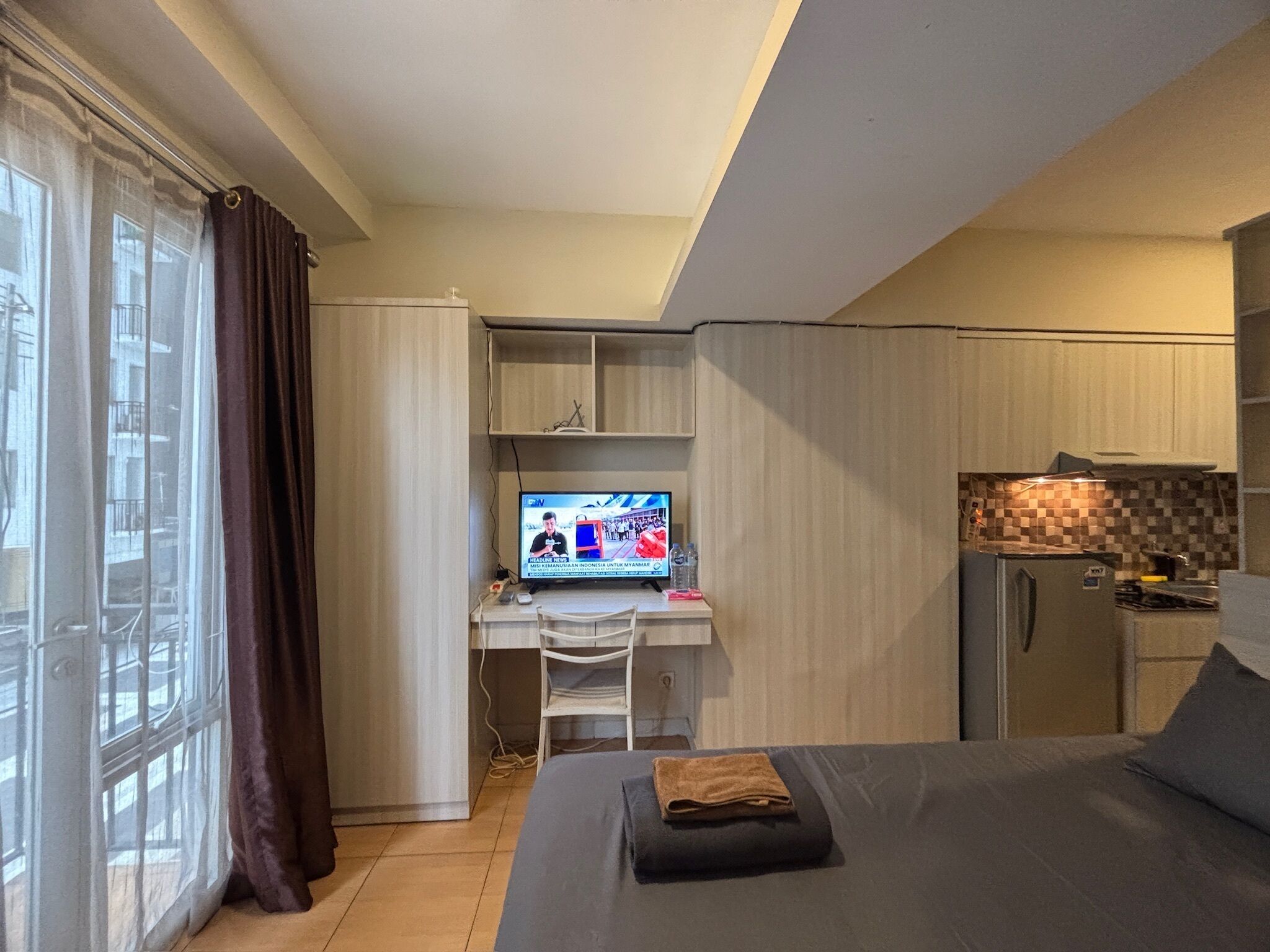 Habitación de hotel pequeña con cama, escritorio con ordenador, refrigerador y ventana con cortinas.