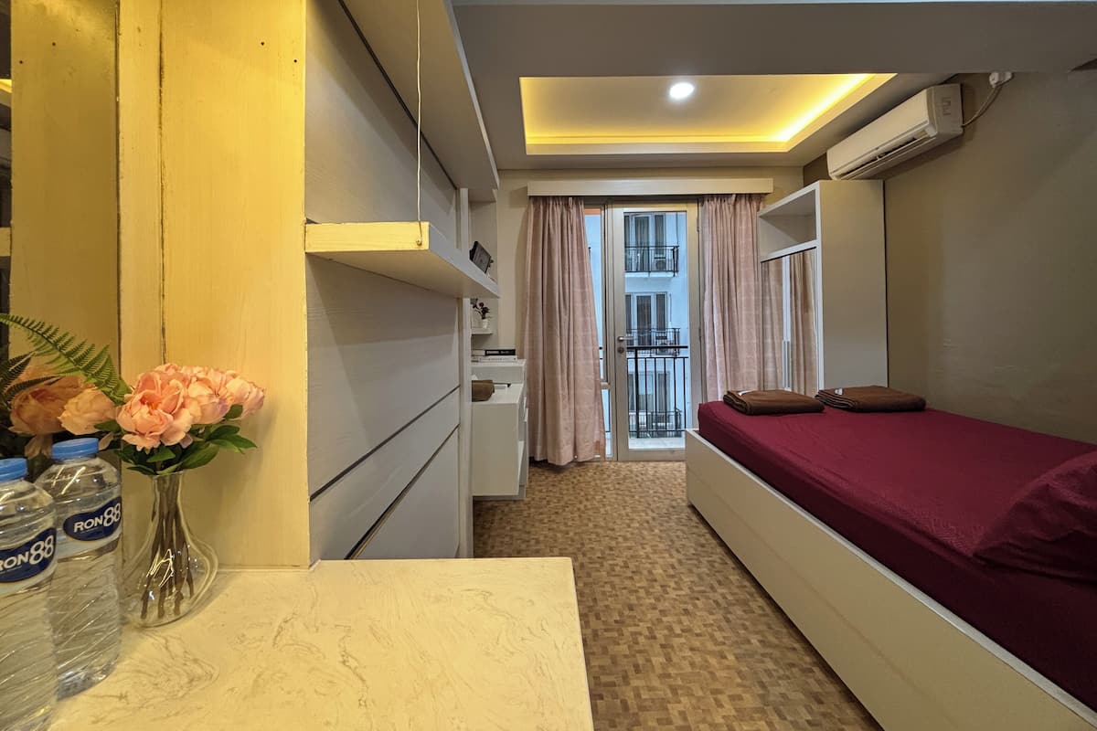 Une chambre d’hôtel de taille réduite, comprenant un lit simple, un petit bureau, une chaise, une fenêtre avec des rideaux, un radiateur et un climatiseur fixé au mur.