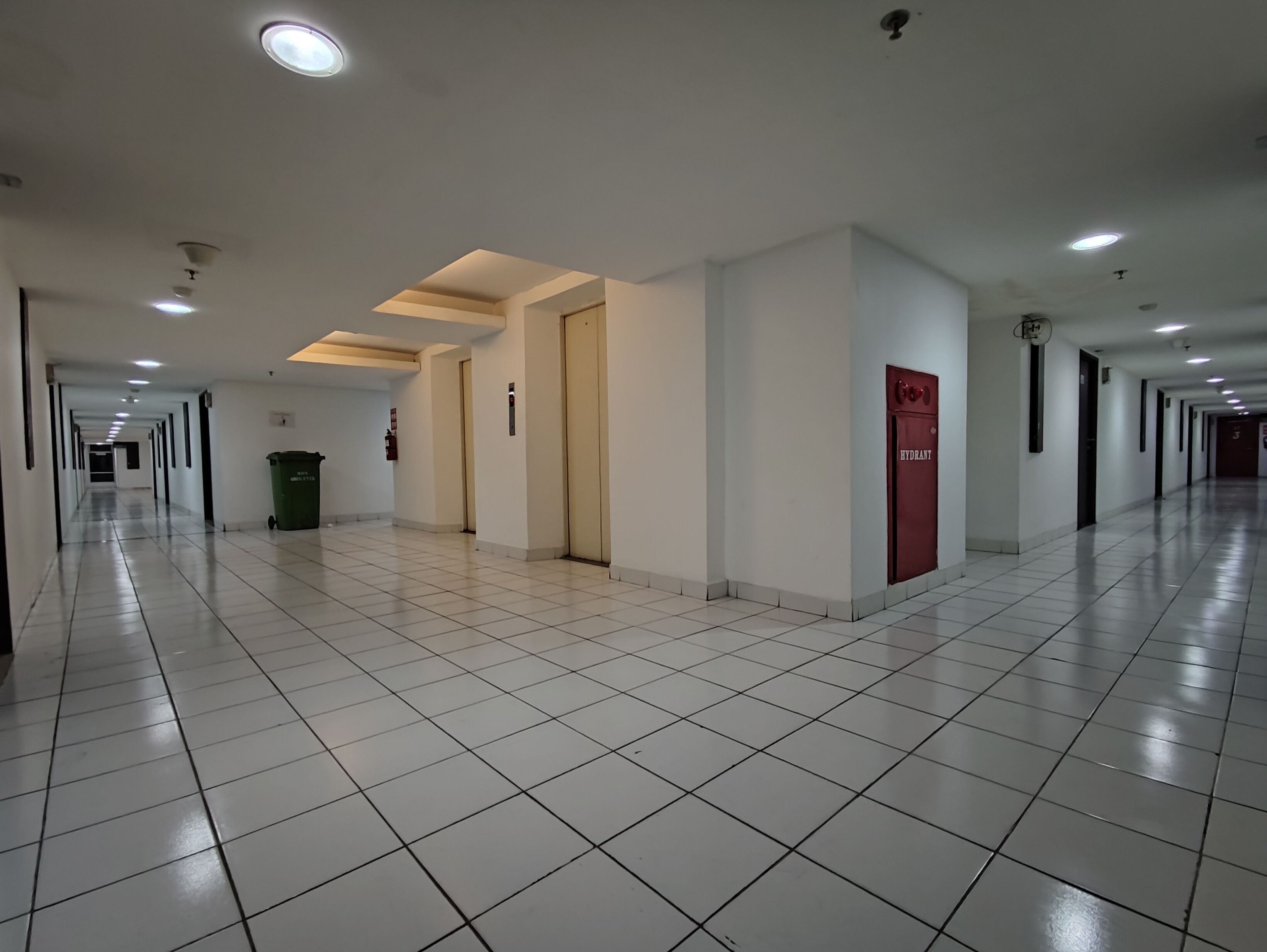 Un long couloir bien éclairé, avec des carreaux au sol, des portes et un poteau d’incendie.