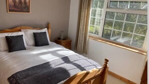 Ein ordentlich bezogenes Bett mit Holz-Kopfstück, ein Nachttisch mit Lampe und ein Fenster mit Vorhängen.