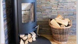 Ein Kaminofen mit schwarzer Außenseite, einem runden Abluftventil und einem Holzkorb daneben.