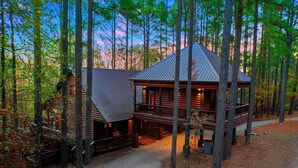 Une cabane en rondins avec un porche, entourée de grands arbres et bordée d’un chemin de gravier.