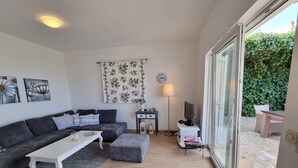 En moderne stue med en grå sofa, et hvidt sofabord og et TV-møbel. Et mønstret gardin, en gulvlampe og udsigt til en have gennem en glasdør.