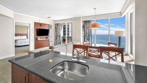 Een moderne hotelkamer met een kitchenette, een eettafel met stoelen en een groot raam met uitzicht op de oceaan.