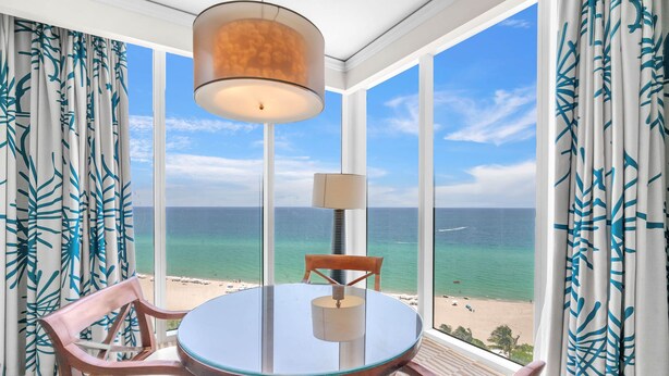Een kamer met een ronde glazen tafel, twee stoelen, een lamp en uitzicht op het strand en de oceaan door grote ramen.