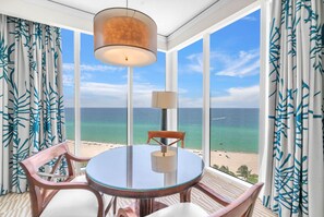 Une pièce avec une table ronde en verre, deux chaises, une lampe et une vue sur la plage et l’océan, grâce à de grandes fenêtres.