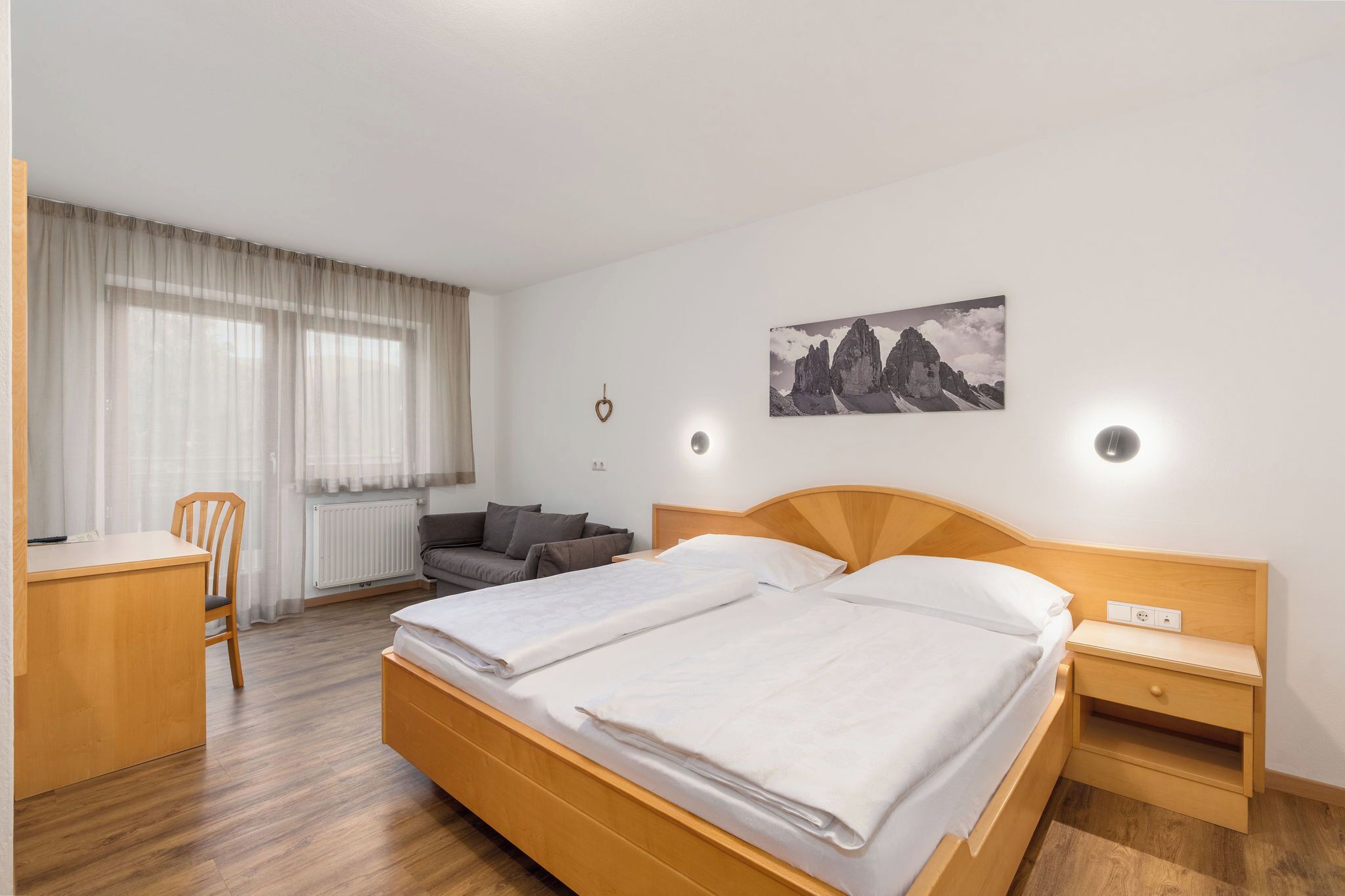 Hotelkamer met een tweepersoonsbed, bureau, stoel, klein tafeltje, nachtkastje en een schilderij aan de muur.