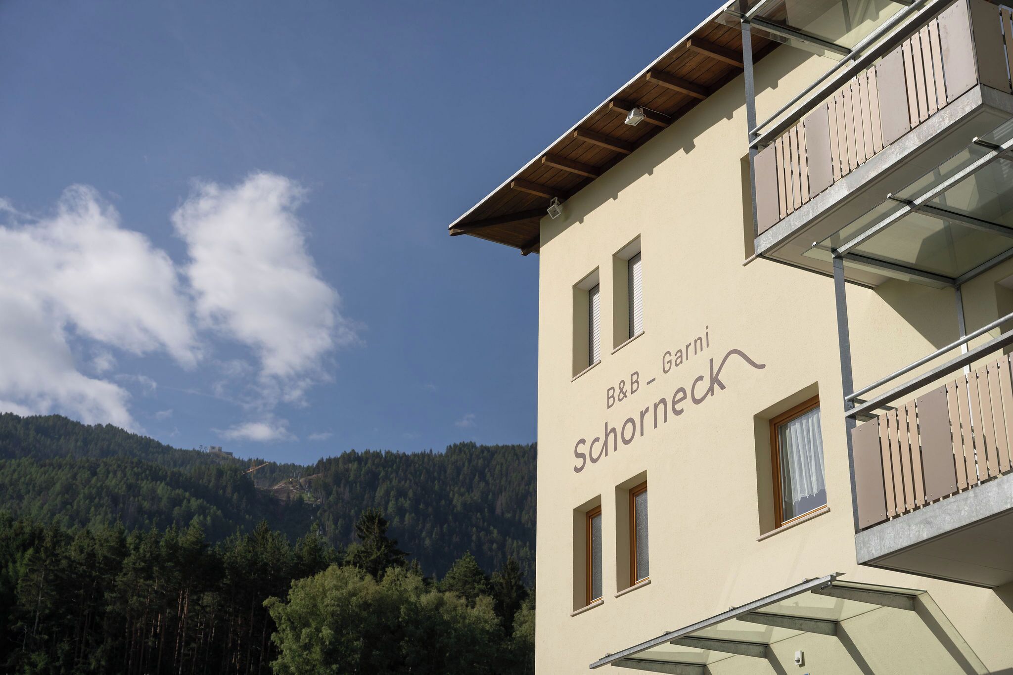 A building with a sign reading 'B&B - Garni Schornerck' and a view of a forested mountain.