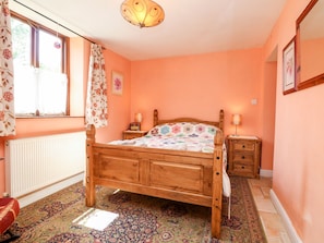 A bedroom with a wooden bed, a nightstand, a lamp, a radiator, and a window with curtains. - The Old Forge Cottage (Bude)