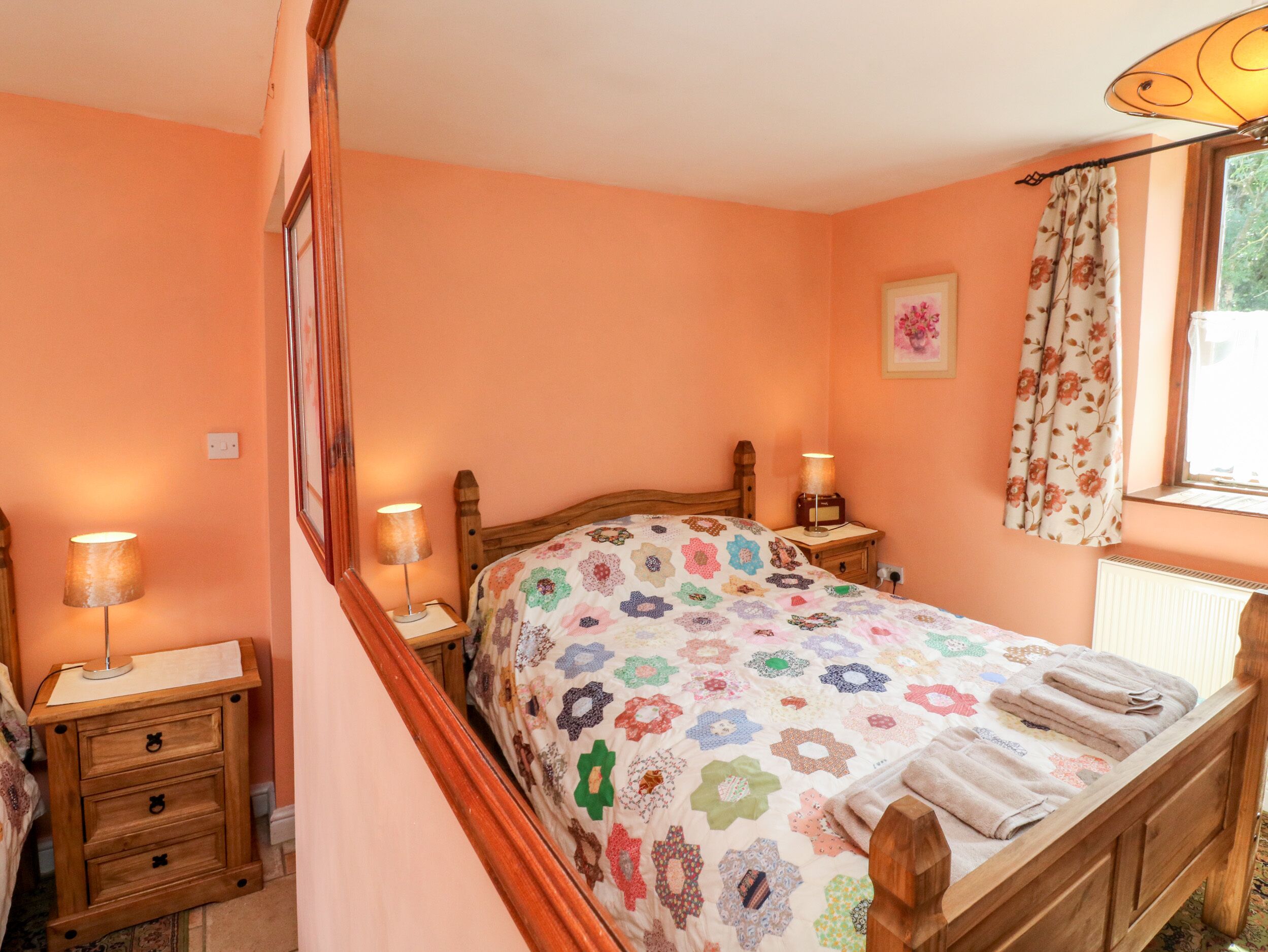 Een slaapkamer met een houten bed, beddengoed met een bloemenprint, bedlampen, een nachtkastje en een raam met gordijnen.