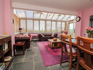 A spacious living room with a large window, leather sofas, a wooden dining table, and a bookshelf. - The Old Forge Cottage (Bude)