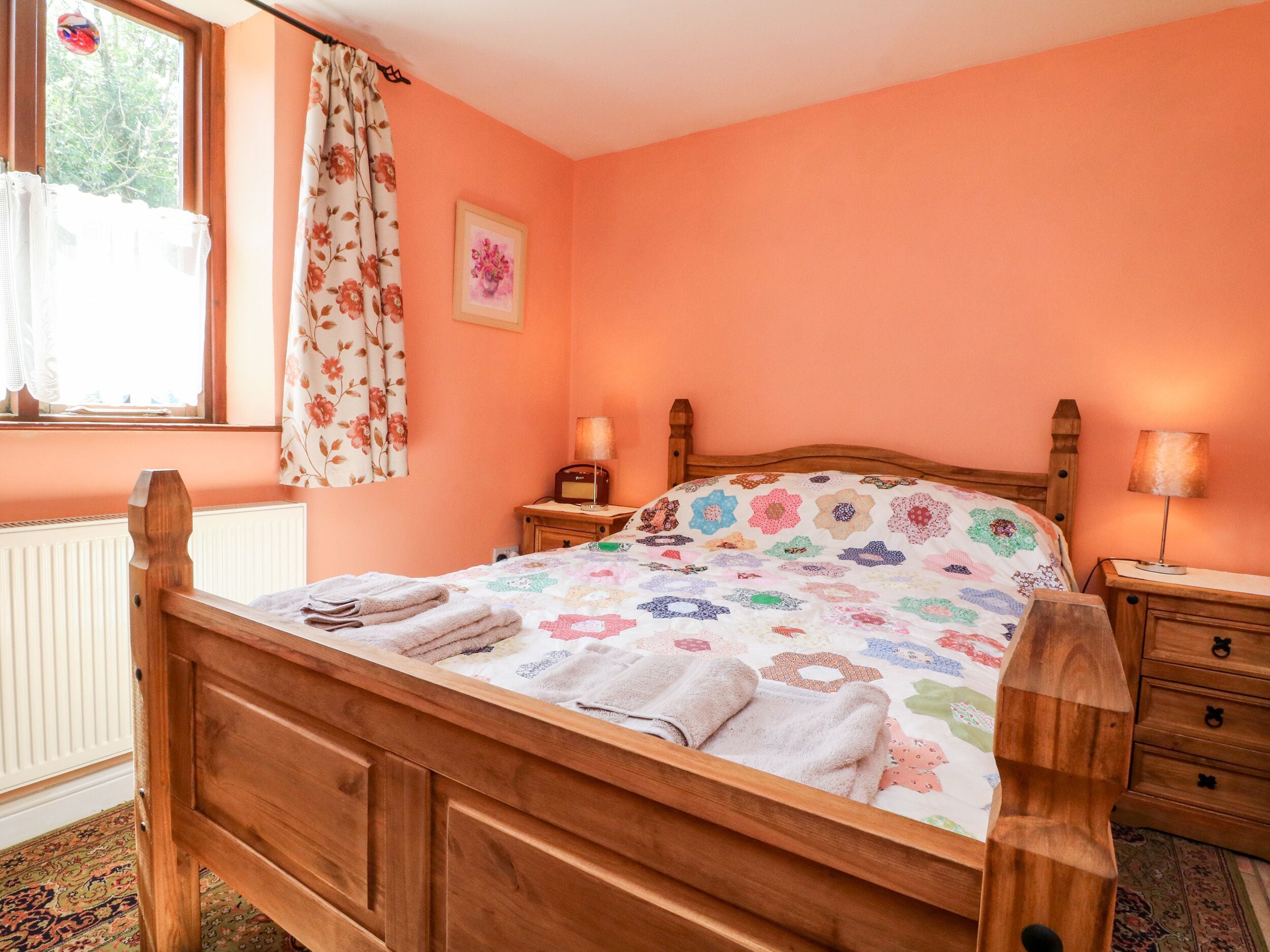 Un dormitorio con una cama de madera, una colcha de estampado floral, una mesita de noche con lámpara y una ventana con cortinas.