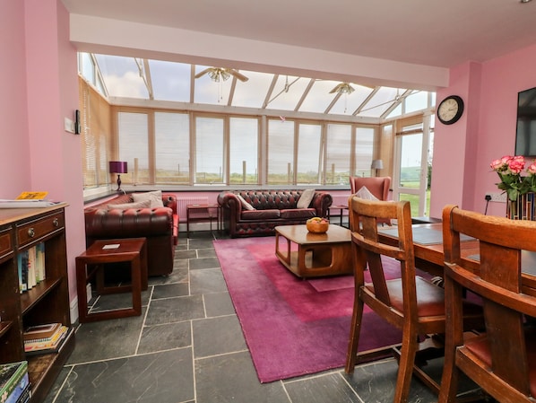 A cozy living room with a fireplace, a large window, and a dining area. - The Old Forge Cottage (Bude)