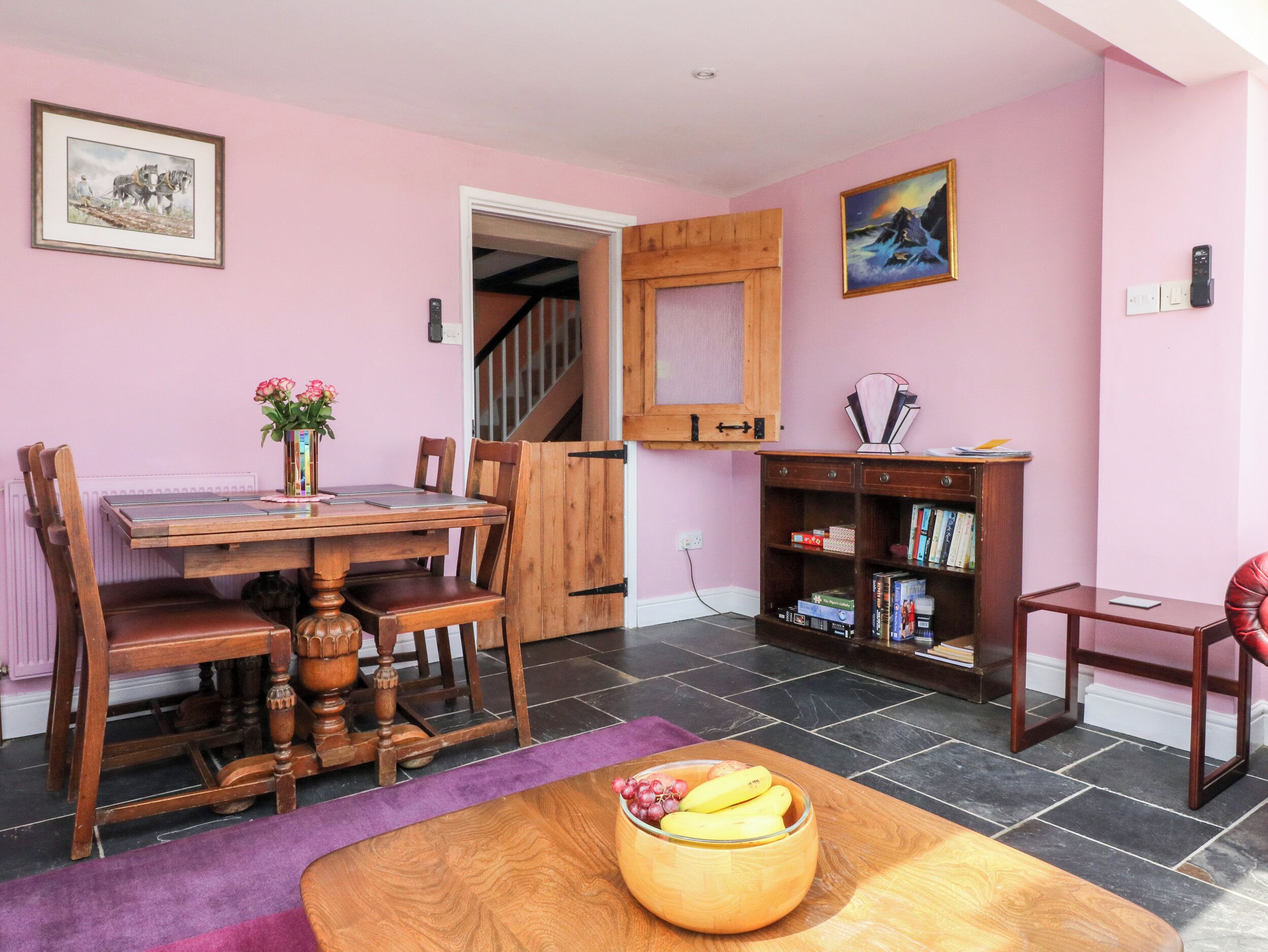 Un comedor con una mesa y sillas de madera, un frutero sobre la mesa, una estantería y una escalera que lleva a otra habitación.