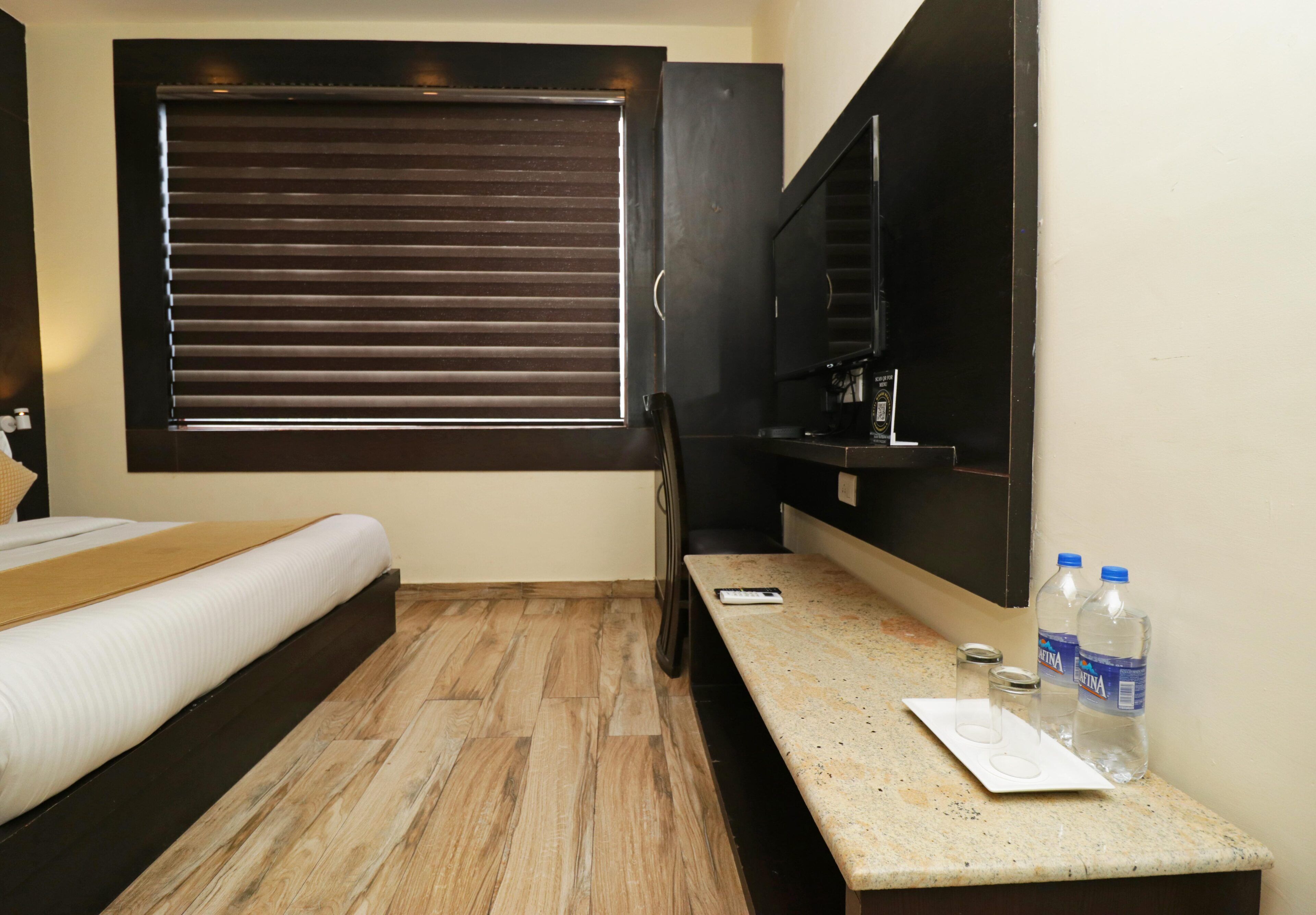 A hotel room with a bed, a desk with a computer, a window with blinds, and a water dispenser.