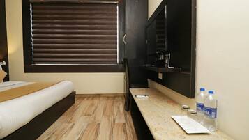 A hotel room with a bed, a desk with a computer, a window with blinds, and a water dispenser.