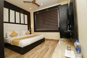 A hotel room with a bed, a ceiling fan, a window with blinds, a flat-screen TV, and a counter with a water bottle and glasses. - Hotel Mirage Residency (Dharamshala)