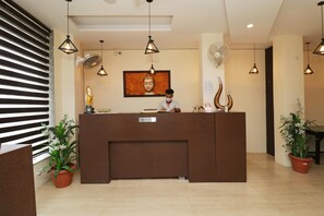 A reception desk with a sign, a Buddha statue, a fan, and potted plants. - Hotel Mirage Residency (Dharamshala)