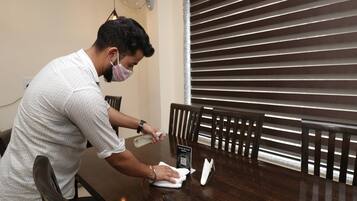 Una persona limpiando una mesa de madera en una habitación con un ventanal grande y un ventilador en la pared.