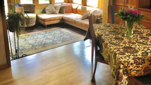 Un salon spacieux doté d’un plancher en bois, d’un canapé avec des coussins, d’une table à manger recouverte d’une nappe à motifs floraux et de grandes fenêtres laissant entrer la lumière naturelle.