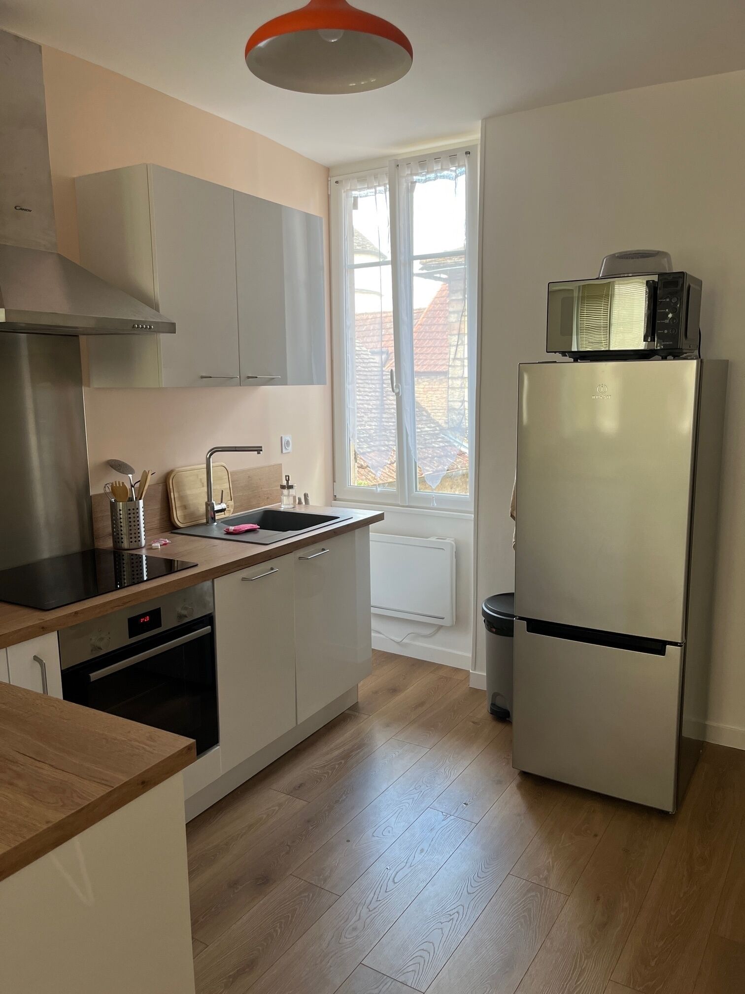 Een moderne keuken met een roestvrijstalen koelkast, een magnetron erboven, houten vloeren en een raam met gordijnen.