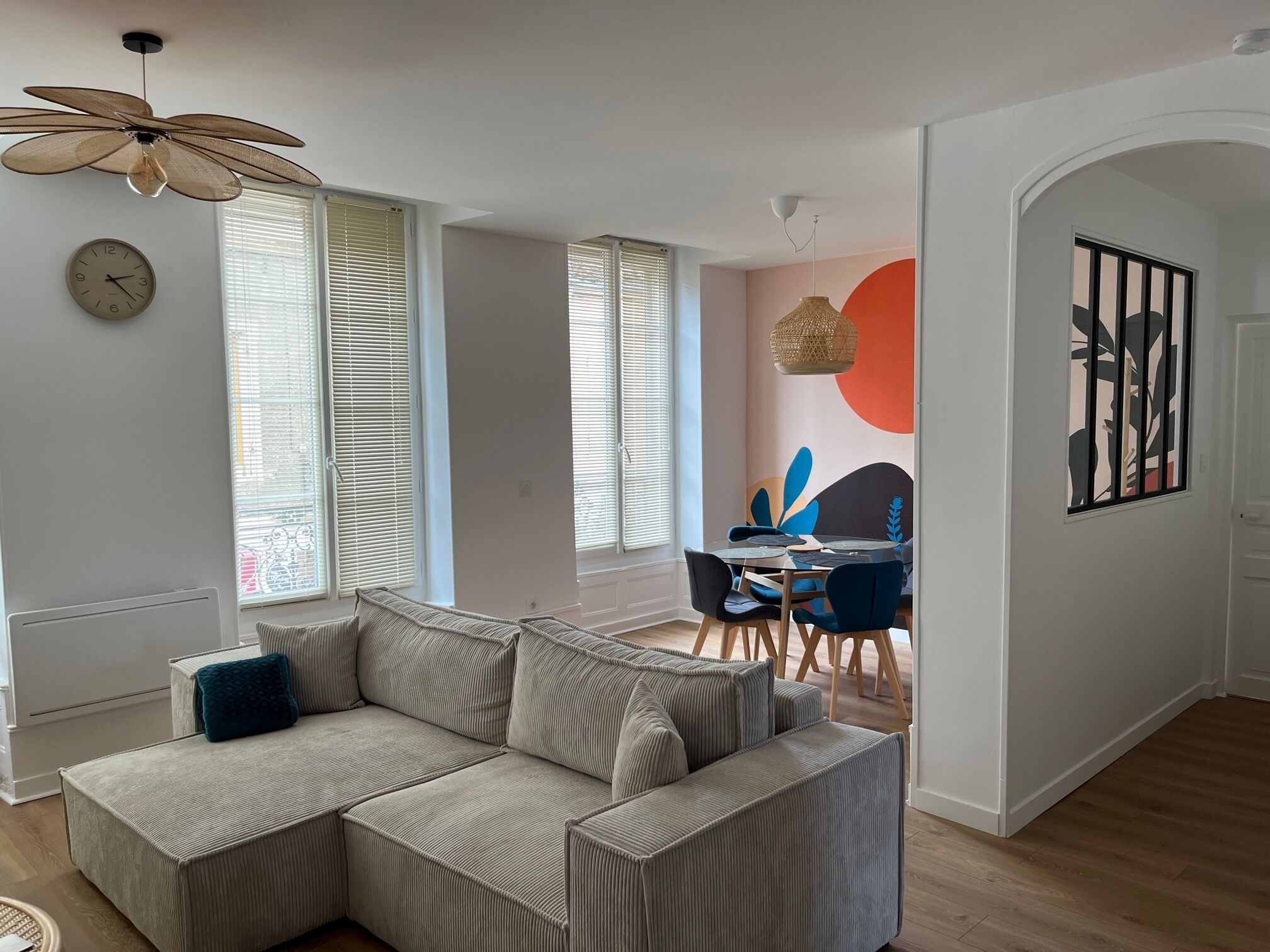 Una sala de estar con un sofá modular, una mesa de comedor con sillas, un ventilador de techo y un reloj de pared.