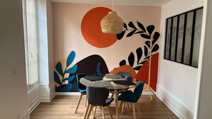 Un coin repas comprenant une table ronde, quatre chaises, un luminaire suspendu en fibres tressées et une fresque murale ornée de motifs abstraits de feuilles.