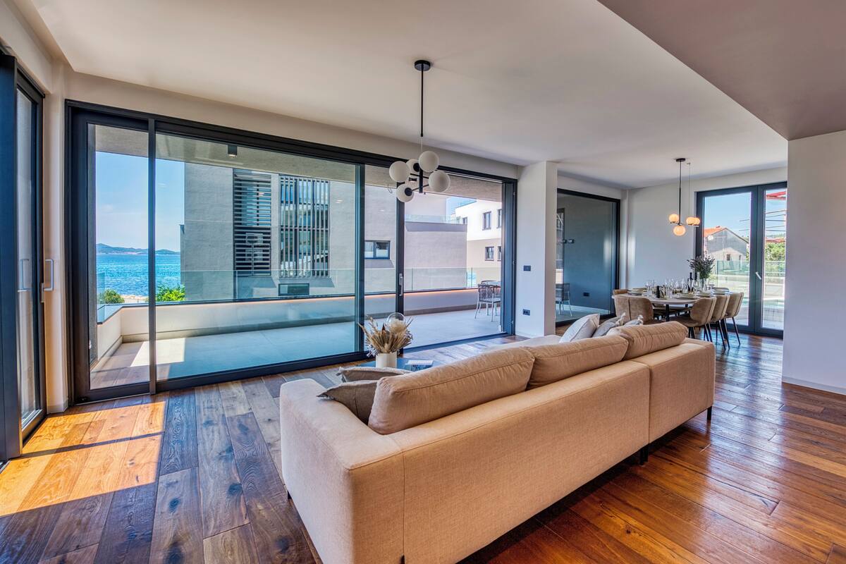 A modern living room with a large sofa, a dining table set for six, and a view of the sea through floor-to-ceiling windows.