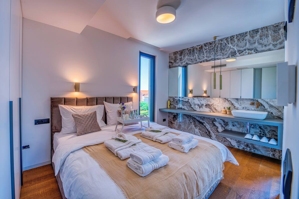 A modern bedroom with a large bed, bedside lamps, a marble bathroom vanity, and a large mirror.