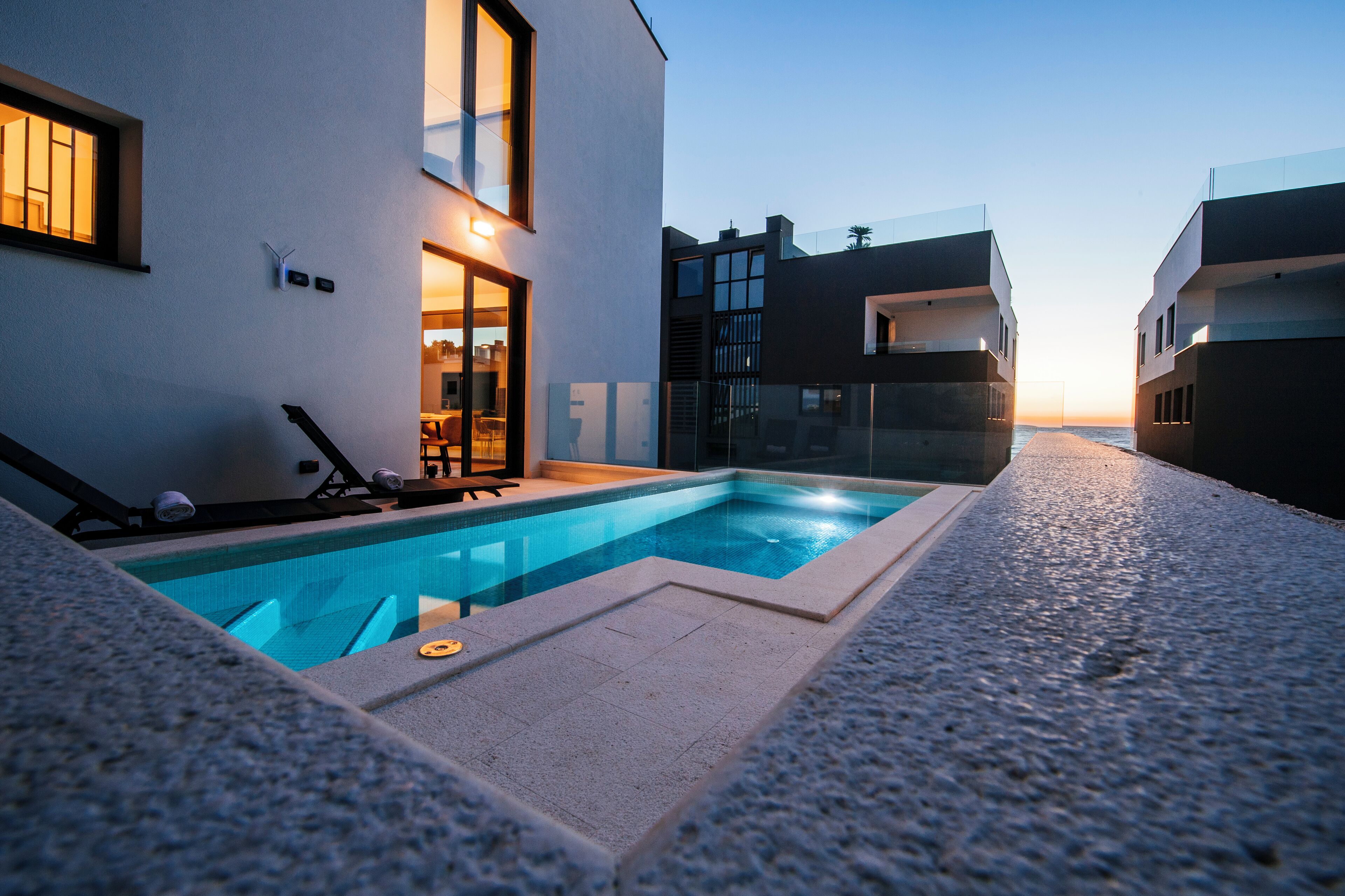 A modern outdoor pool area with a clear view of the sunset, a lounge chair, and a glass-enclosed door leading to a room.