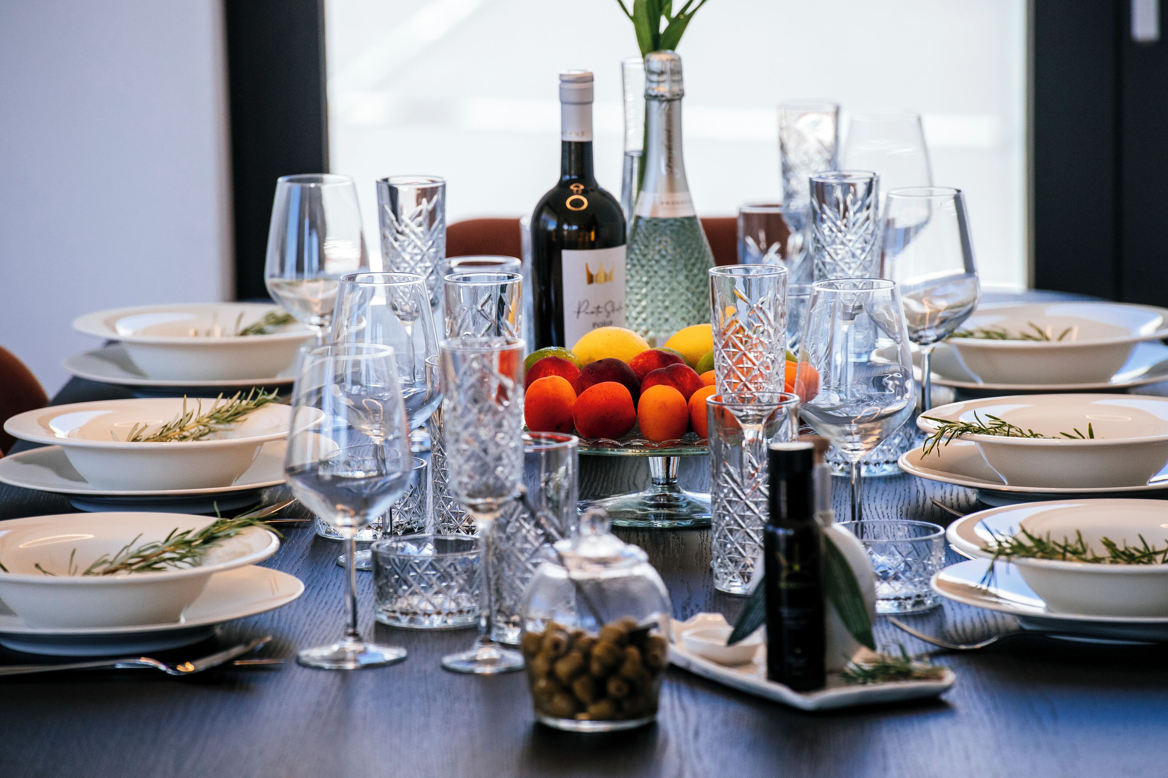 Mesa de jantar posta com vários pratos, copos e uma garrafa de vinho, rodeada por taças decorativas e frutas frescas.