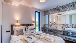 A modern bedroom with a large bed, bedside lamps, a marble bathroom vanity, and a large mirror.
