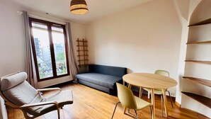 A modern living room with a sofa, a dining table with chairs, a hanging light fixture, and a view of buildings through large windows. - Appartement Ménilmontant (Paris)