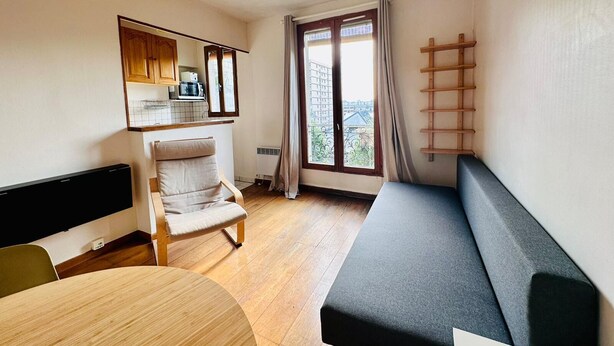 A compact living space with a kitchenette, a dining area, a sofa, and a window with a view of buildings. - Appartement Ménilmontant (Paris)