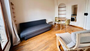 A modern living room with a sofa, a dining table, and a chair. There is a window with curtains, a ceiling light, and a door with a mirror. - Appartement Ménilmontant (Paris)