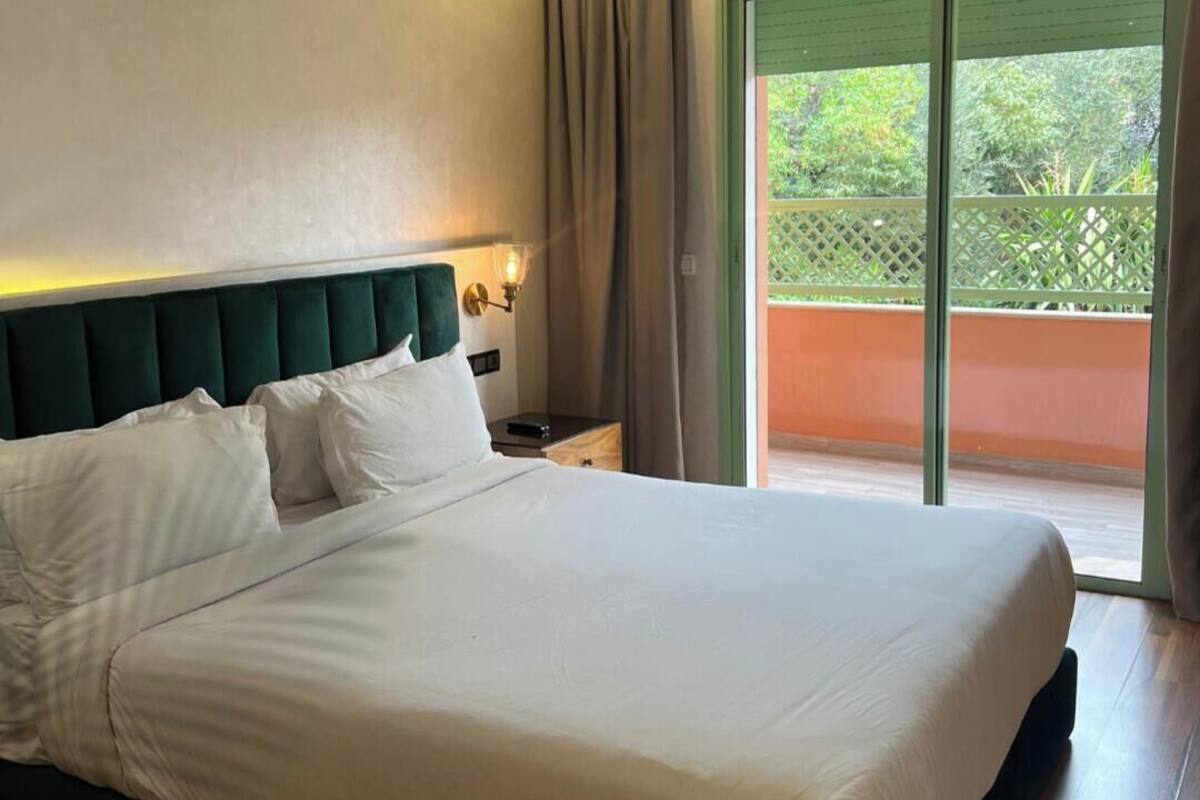 A neatly made bed with white linens in a room with a green upholstered headboard, a nightstand with a lamp, and a sliding glass door leading to an outdoor area.