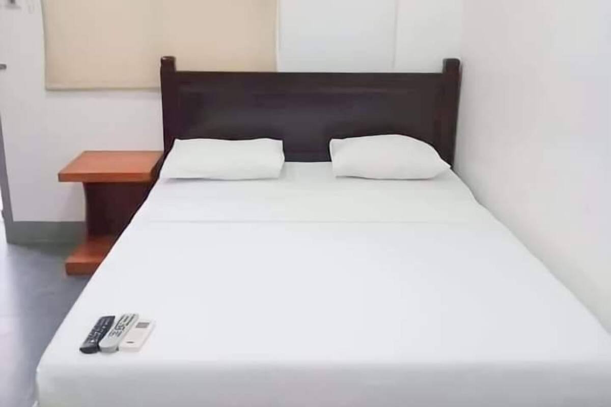 Una cama individual con cabecera de madera, ropa de cama blanca y una mesita de noche con un control remoto.