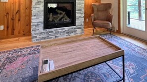 Un salón acogedor con chimenea de piedra, paredes de madera, un televisor montado y un ventanal grande.