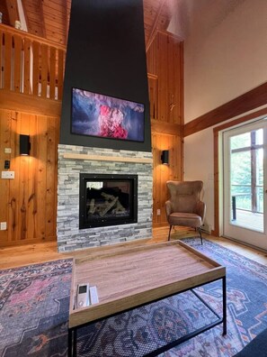 A cozy living room with a stone fireplace, wooden walls, a mounted TV, and a large window. - Hemlock Chalet @ Brickyard Creek, Bayfield (Bayfield)