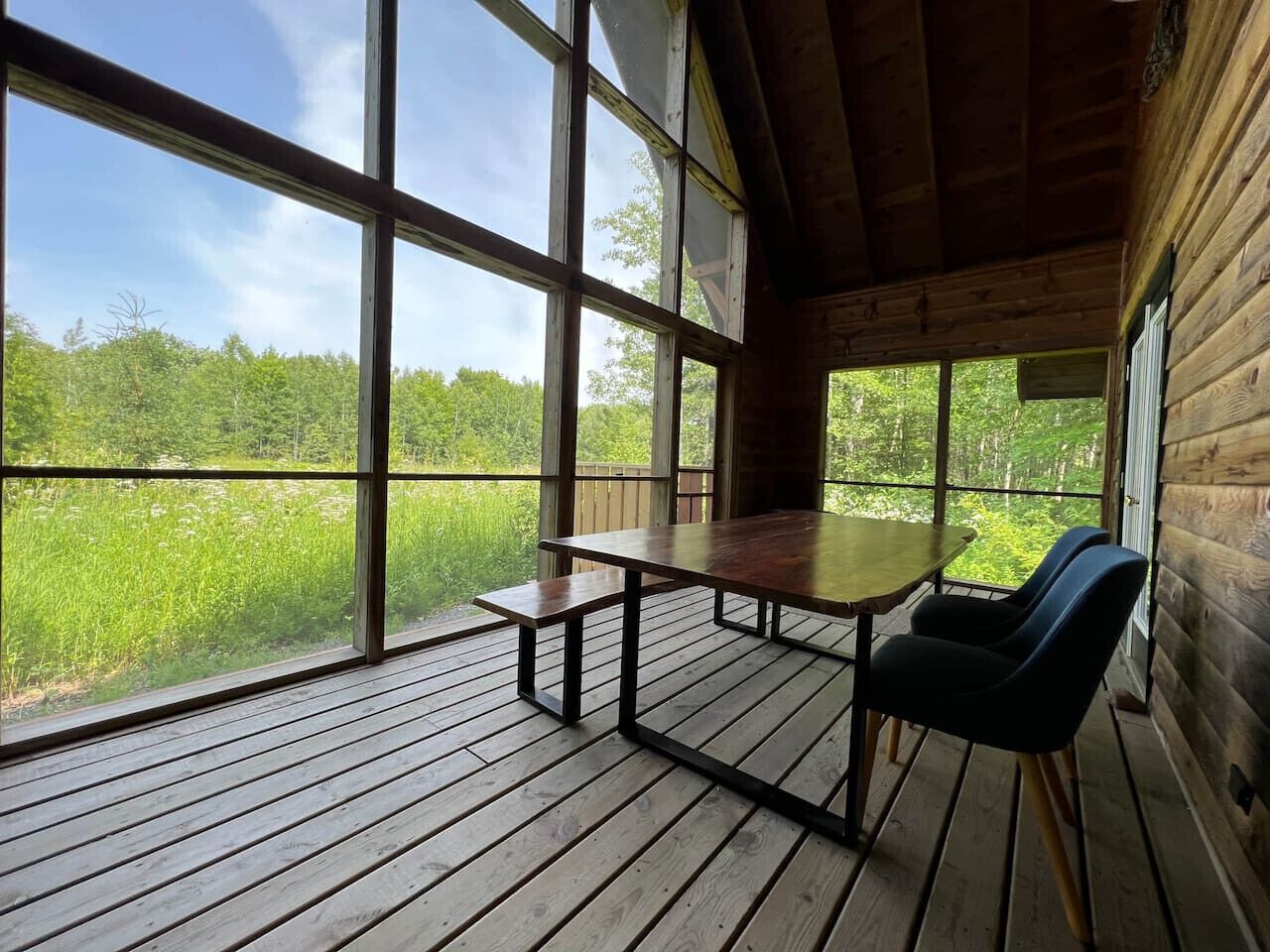 Un patio in legno con tavolo e panchina, ampie finestre che si affacciano su un paesaggio verde.