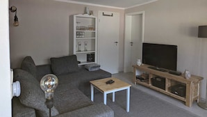 A living room with a grey sofa, a coffee table, a TV on a wooden stand, and a white shelving unit with glassware. - Double vacation bungalow in the middle of the Schorfheide (Joachimsthal)