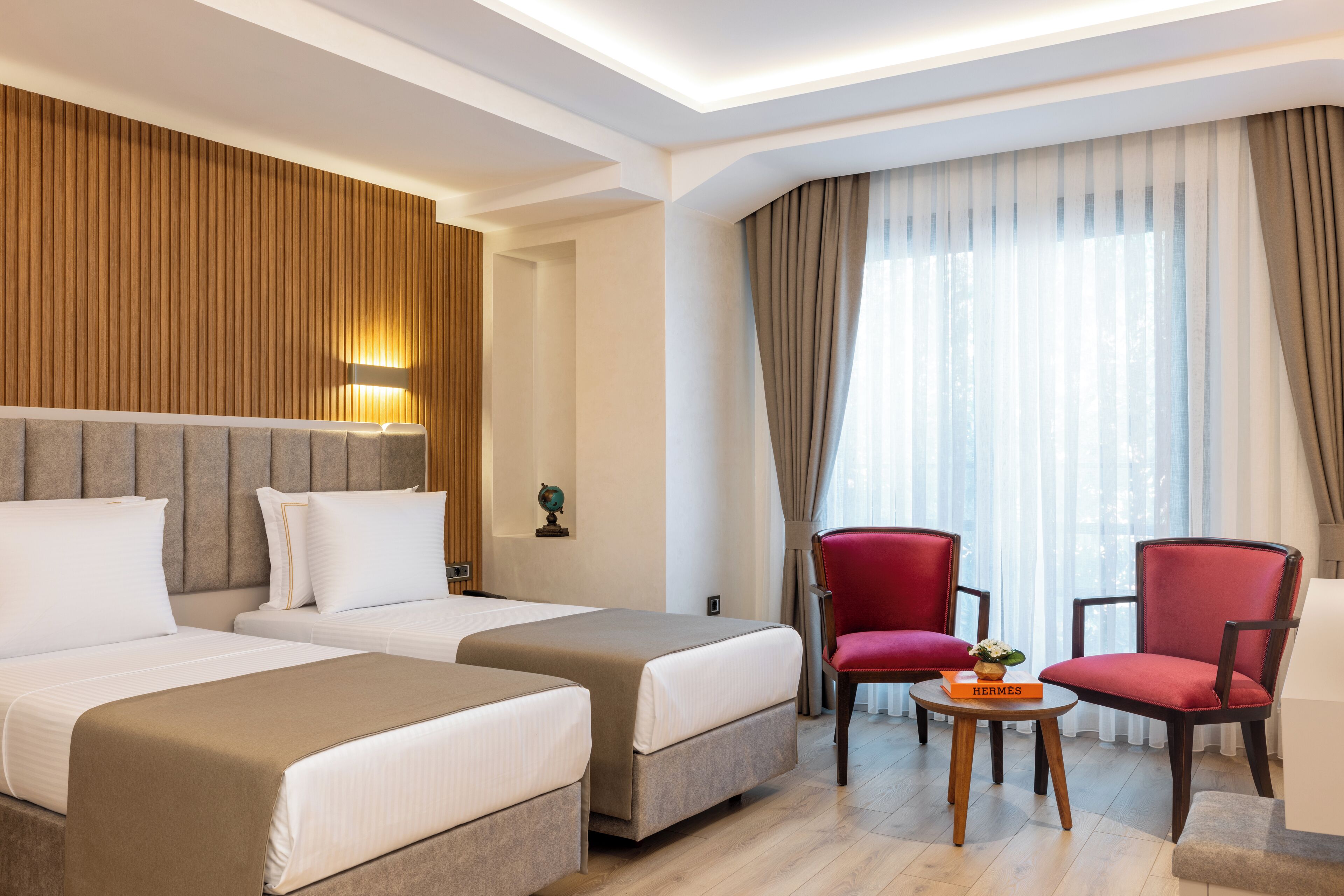 Habitación de hotel con dos camas, un sillón rojo, una mesita con un libro y un ventanal con cortinas.