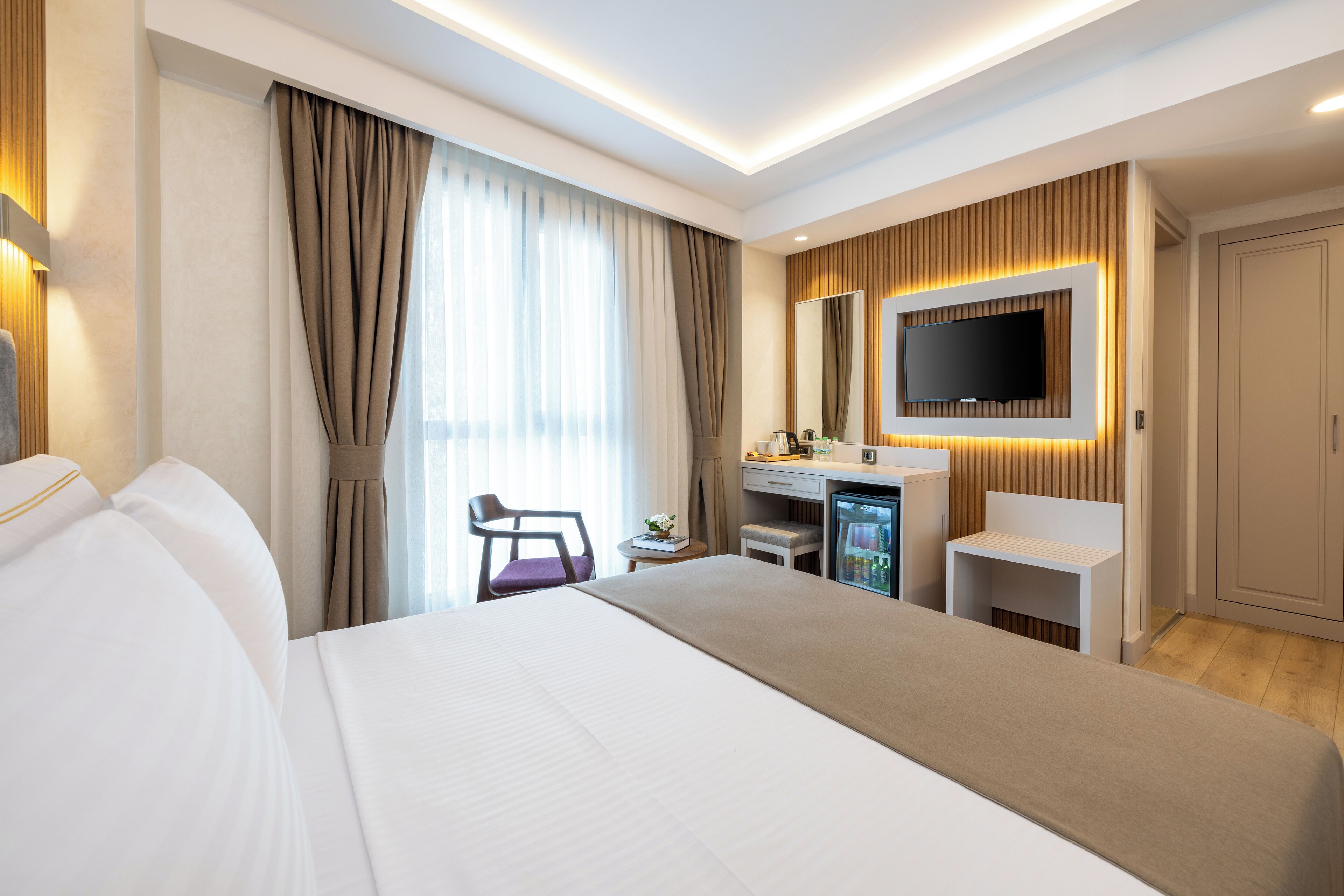 A modern hotel room with a large bed, a desk with a computer, a television, and a minibar.