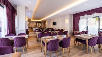 Un comedor amplio con múltiples mesas y sillas tapizadas en morado, con una lámpara moderna y un televisor de pantalla plana montado en la pared.