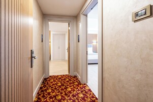 A hotel hallway with a patterned carpet, a door with a handle, and a wall-mounted device.