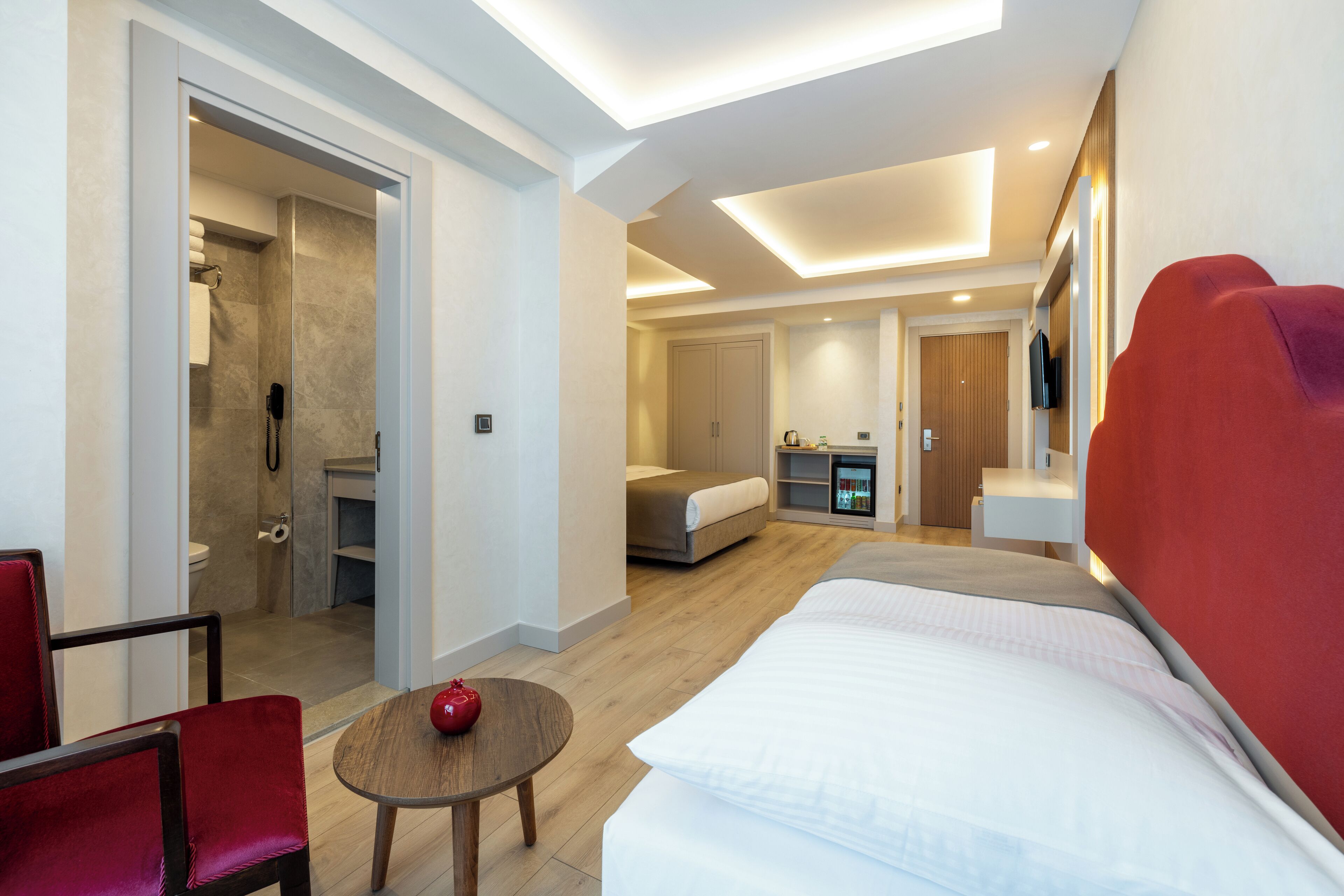 Una habitación de hotel moderna con una cama, una mesita con un objeto rojo, una silla roja, zona de ducha y una kitchenette.