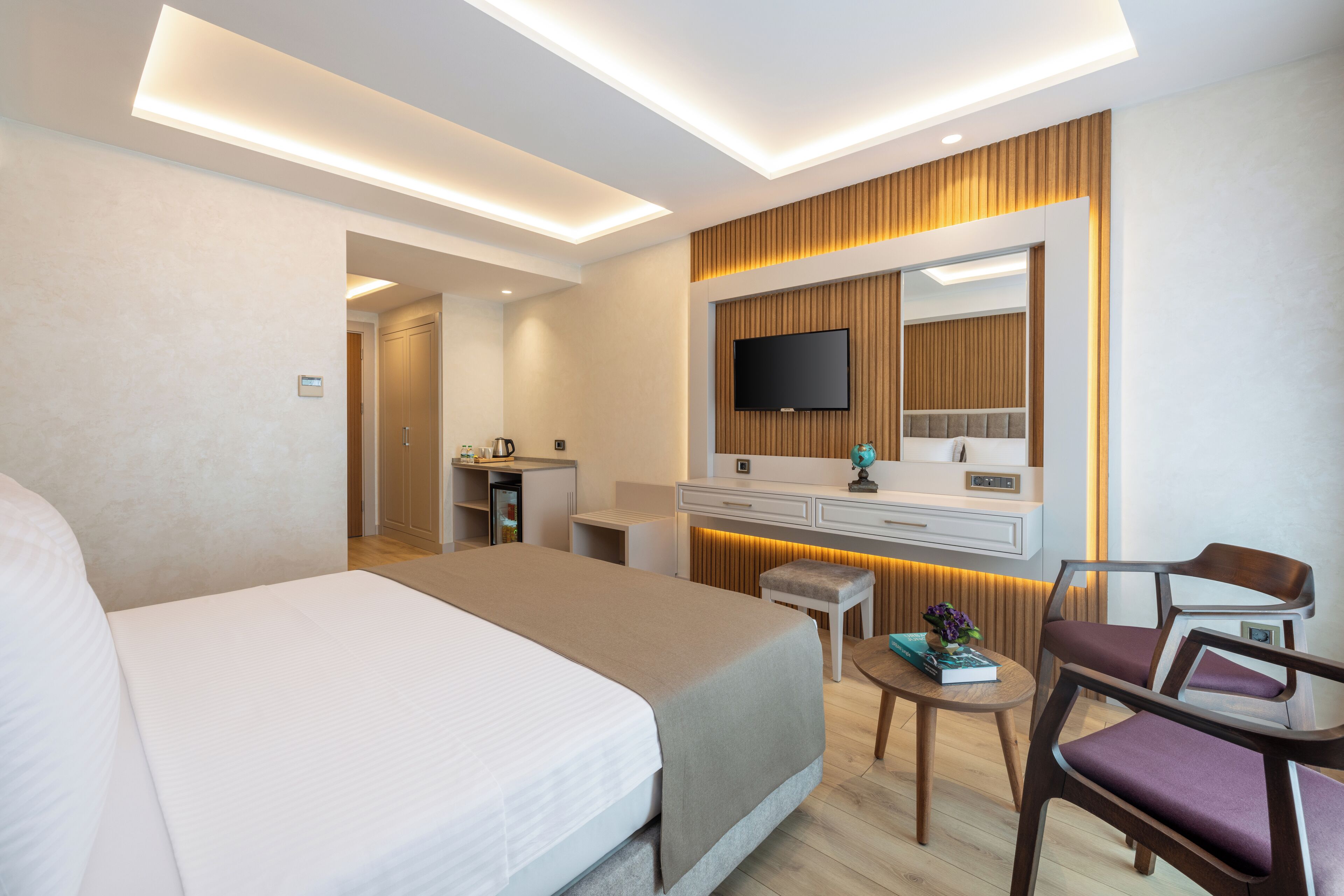 Une chambre d’hôtel moderne avec un grand lit, un bureau, une chaise, une télévision et une petite table sur laquelle se trouvent un livre et une plante.