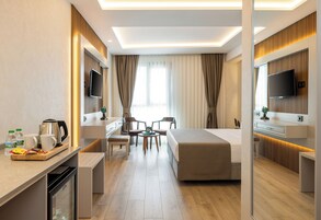 A modern hotel room with a bed, a desk with a coffee maker, a TV, and a seating area with chairs.