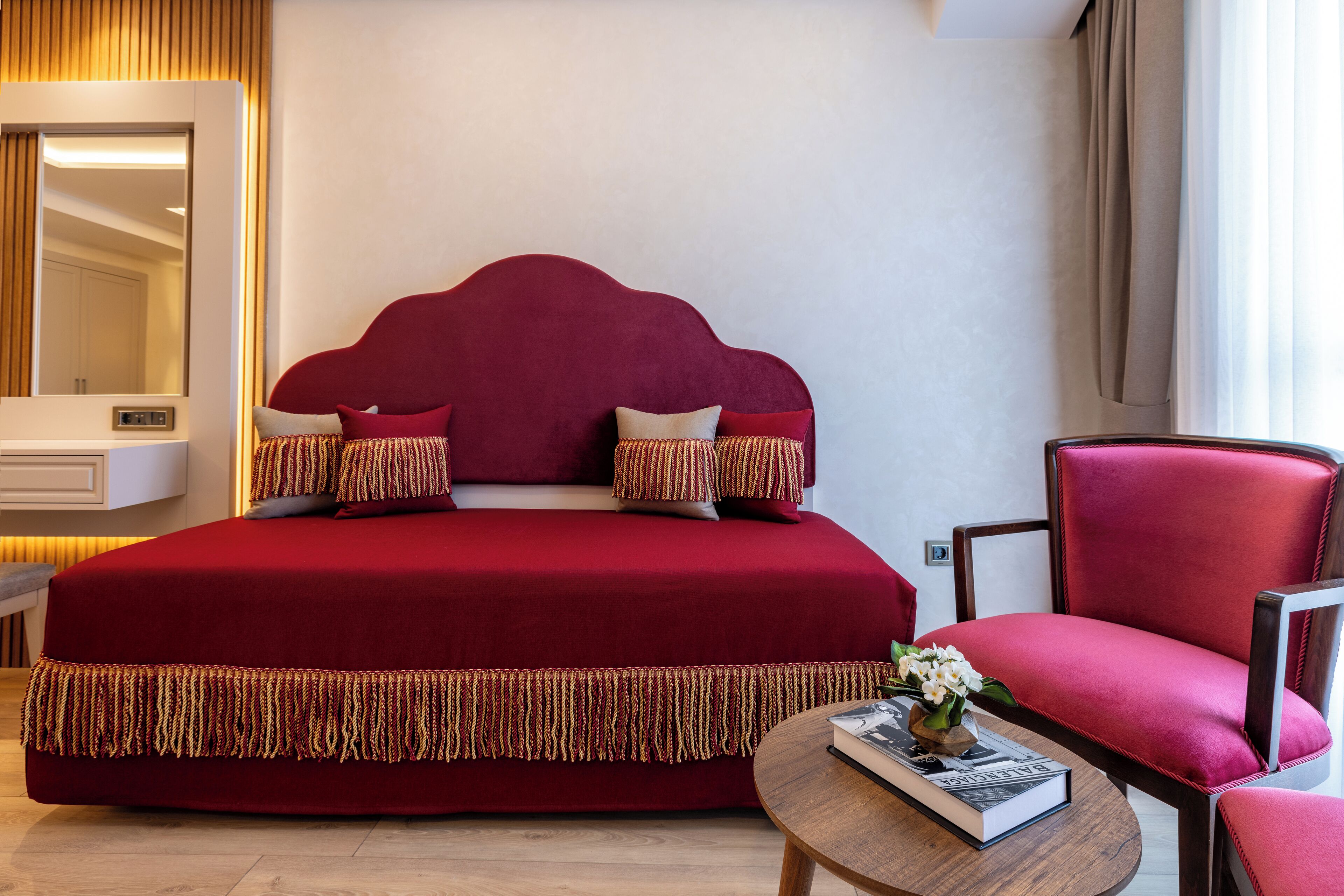 Una cama grande con cabecera roja, un sillón tapizado en rojo, una mesita de noche con un libro y un espejo con marco de madera.