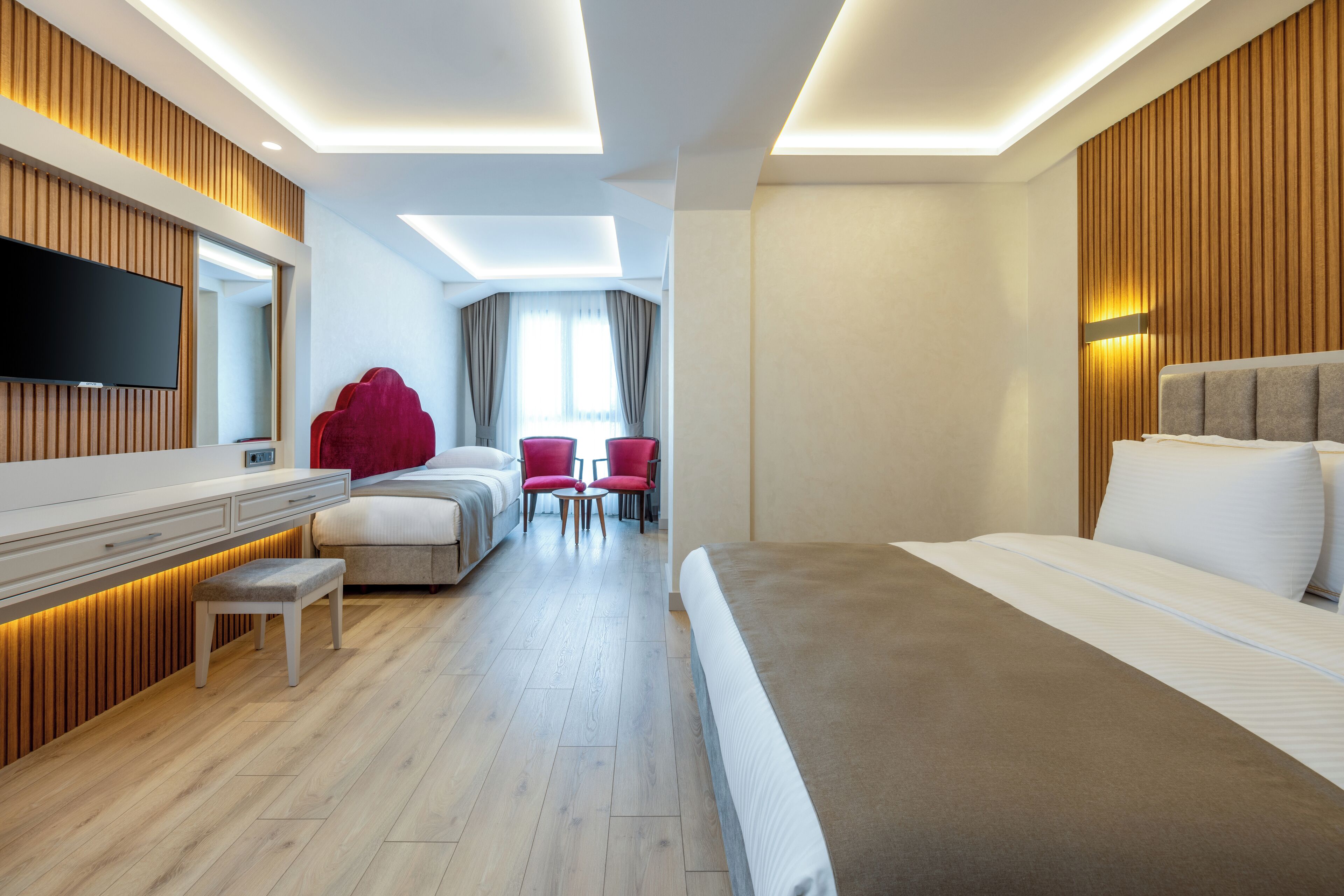 A modern hotel room with a large bed, a flat-screen TV mounted on the wall, a seating area with two chairs, and a wooden accent wall.